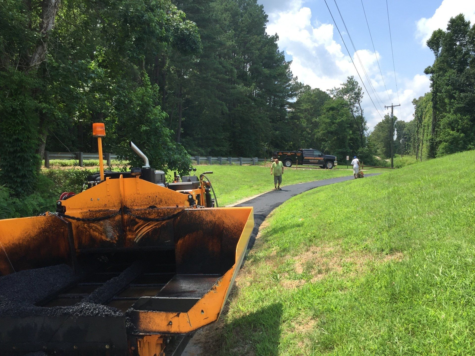 Asphalt paving in progress on a road, with workers, machinery, and trees.