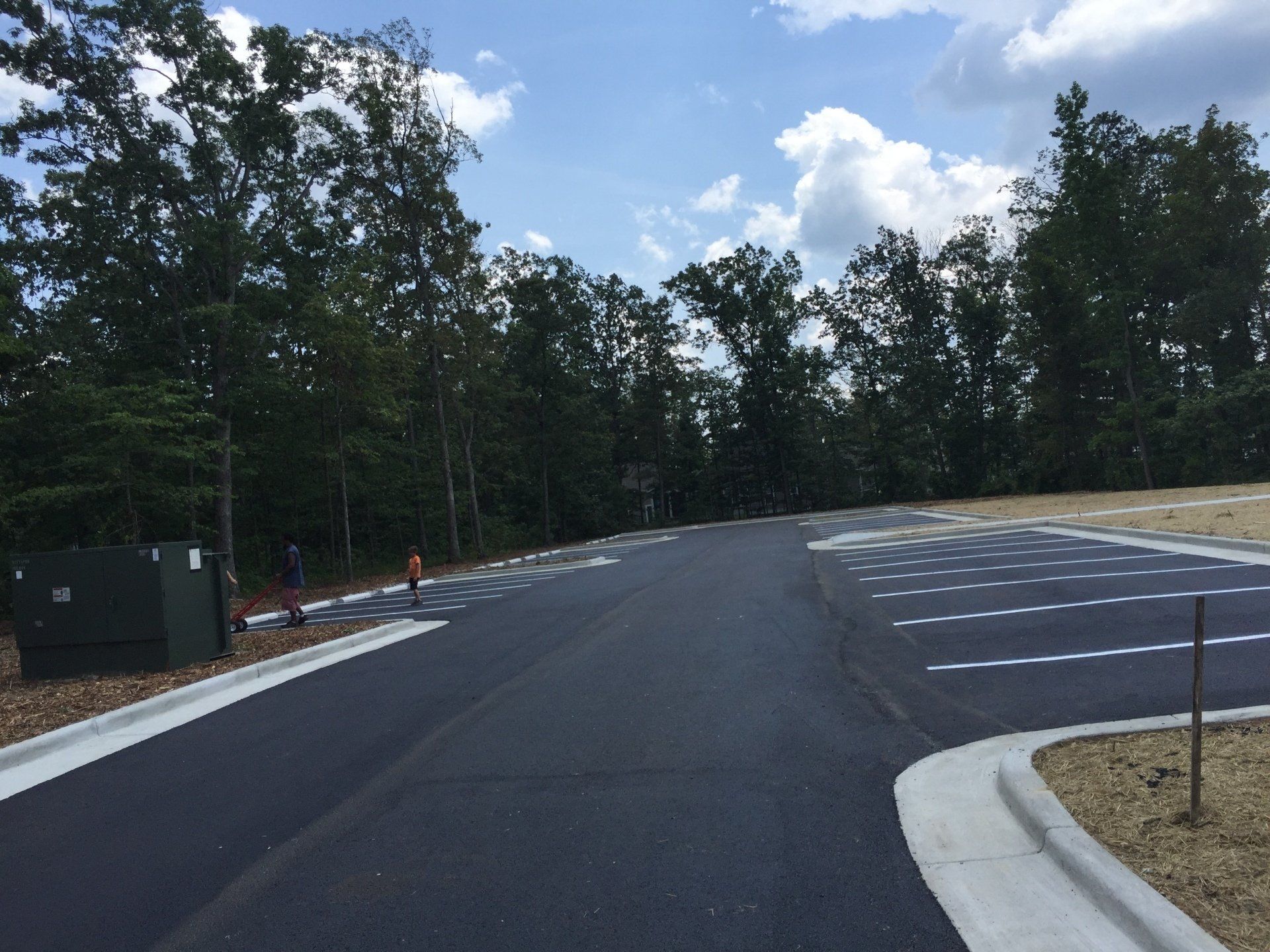Asphalt road with parking spaces, bordered by concrete and trees, against a cloudy sky.