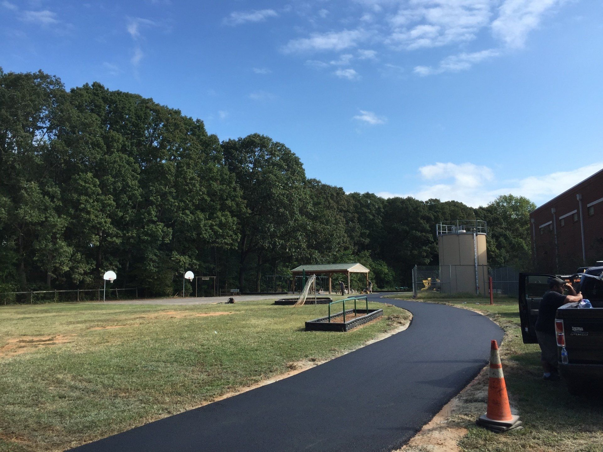A newly paved asphalt path curves through a grassy area, alongside trees and a building on a sunny day.