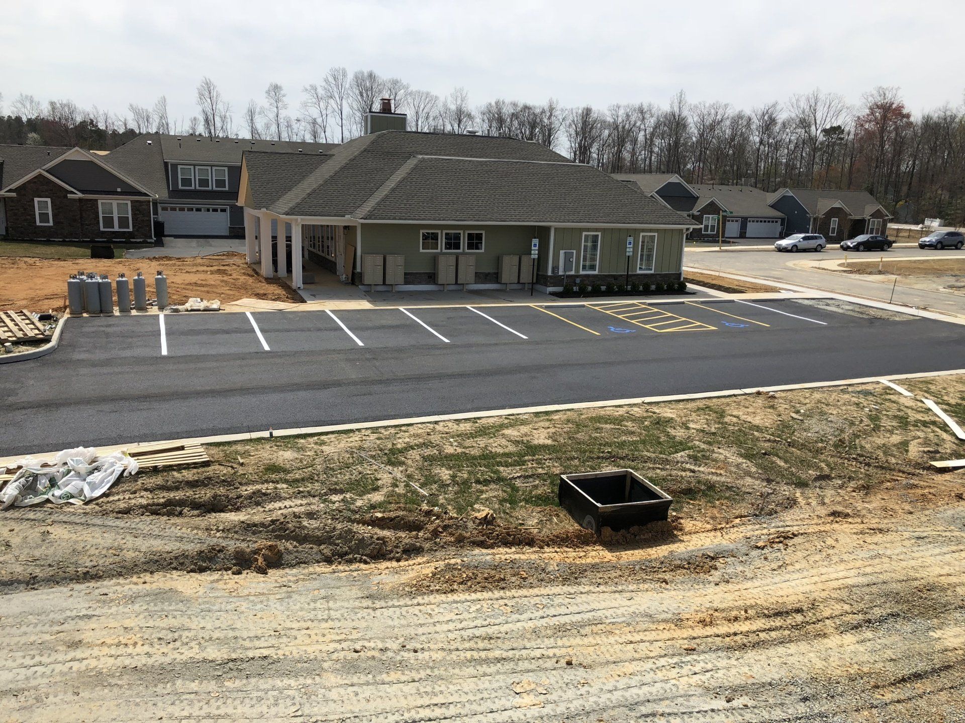 Newly paved parking lot in front of a light green building. Several houses in the background, construction debris visible.