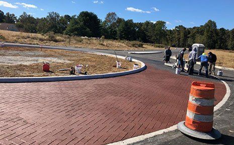 Workers applying reddish-brown coating to new brick-patterned road surface. Orange traffic cone present.