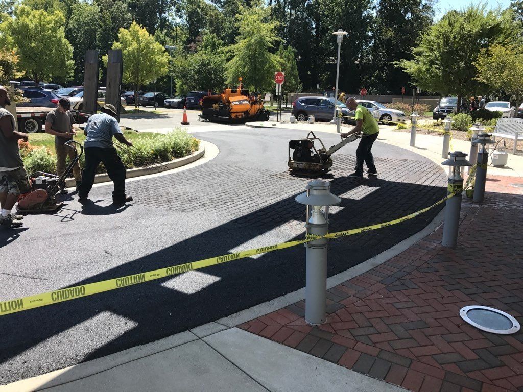Asphalt paving crew at work, applying fresh asphalt to a circular driveway. Yellow caution tape in foreground.