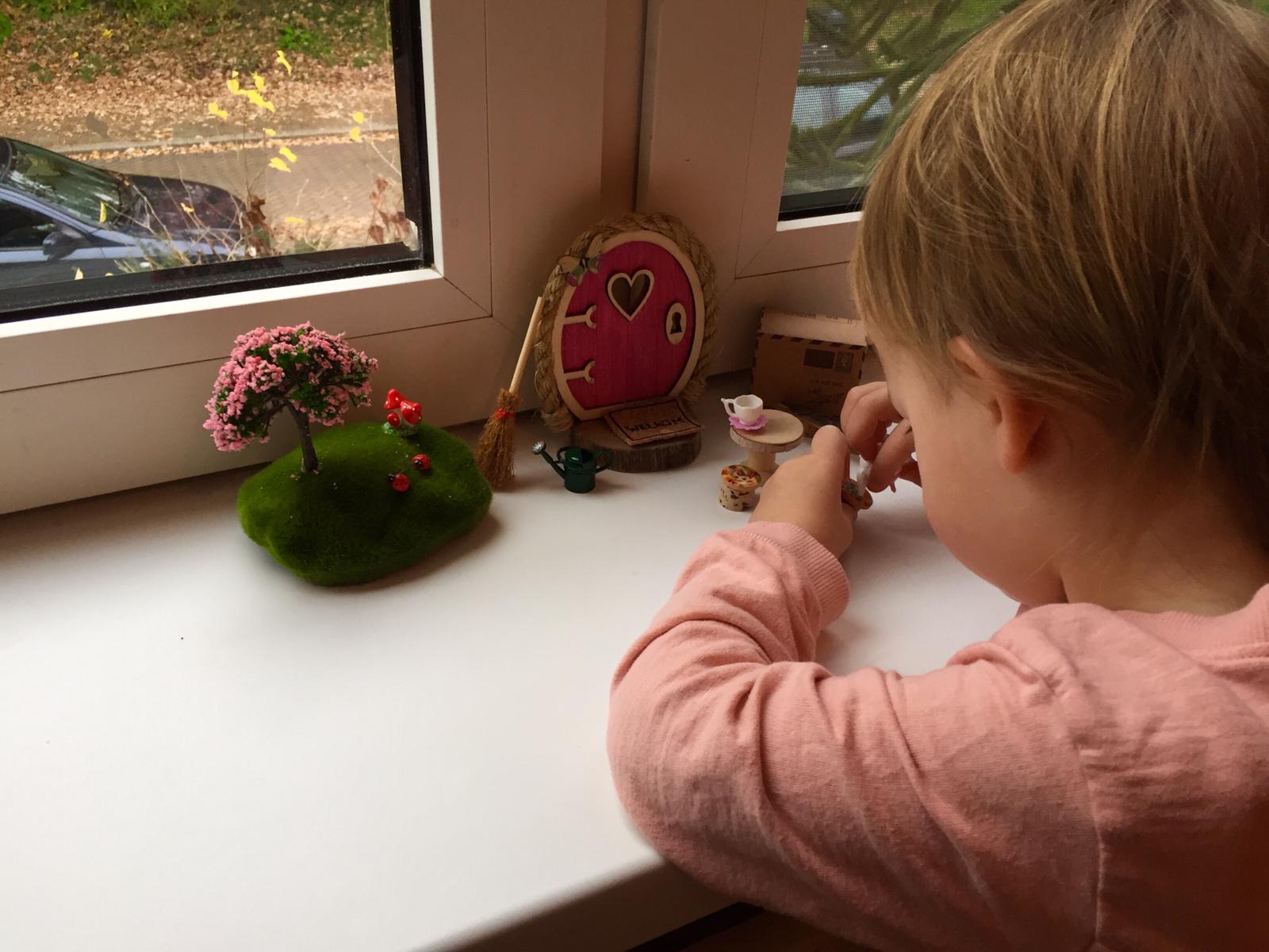 Een kind kijkt naar een miniatuur sprookjestuin op de vensterbank. Inclusief een klein deurtje, boompje en meubeltjes.