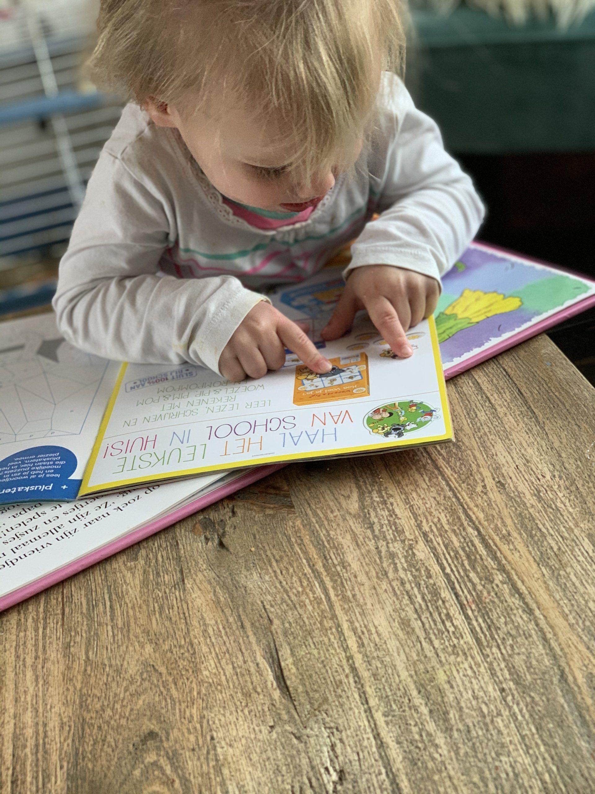 Kind met blond haar leest een boek en wijst met beide wijsvingers naar een plaatje.