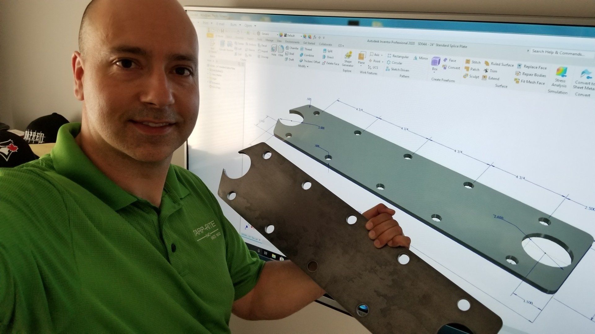 A man in a green shirt is holding a piece of metal in front of a computer screen.