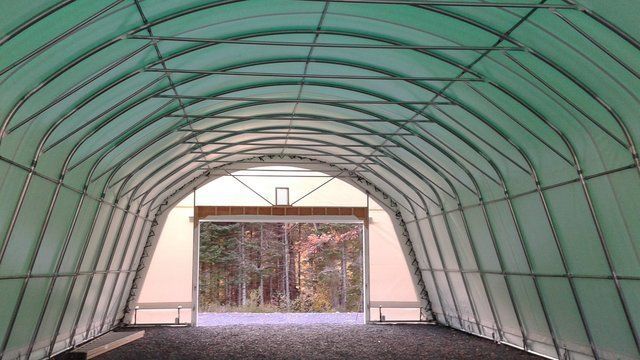 Interior view of a green tarp-covered structure with a doorway opening to trees. Metal frame arches. Cover-Tech Portable Garages.