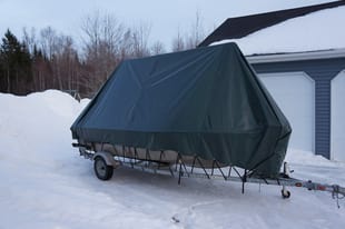 A boat trailer with a tarp on it is parked in the snow in front of a garage.