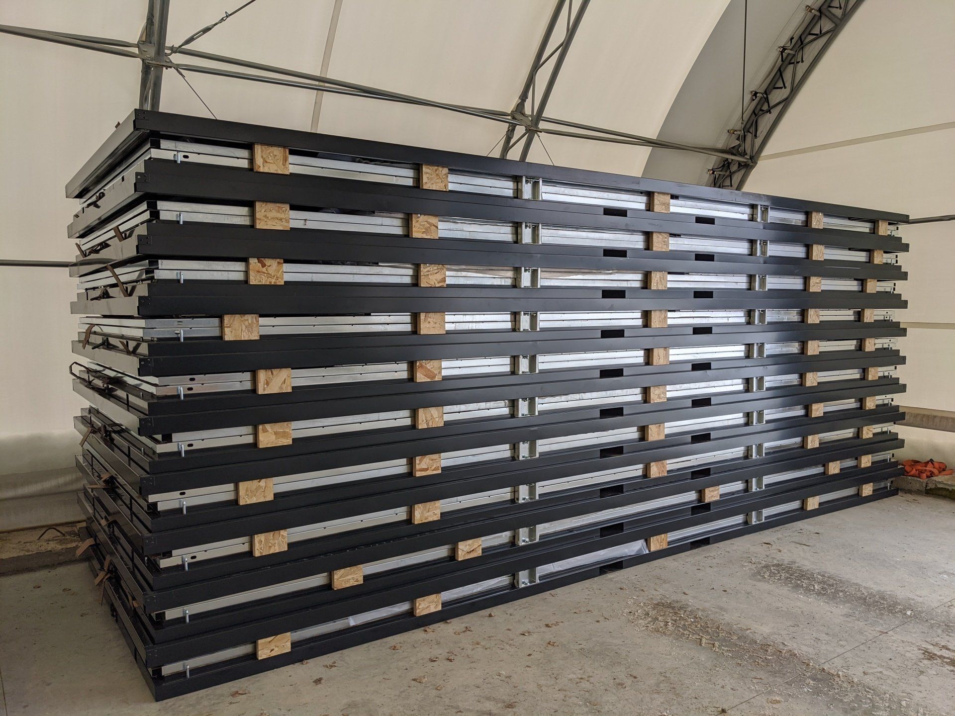 Large stack of dark gray panels, separated by wooden supports, inside a large tent. Cover-Tech Portable Containers.