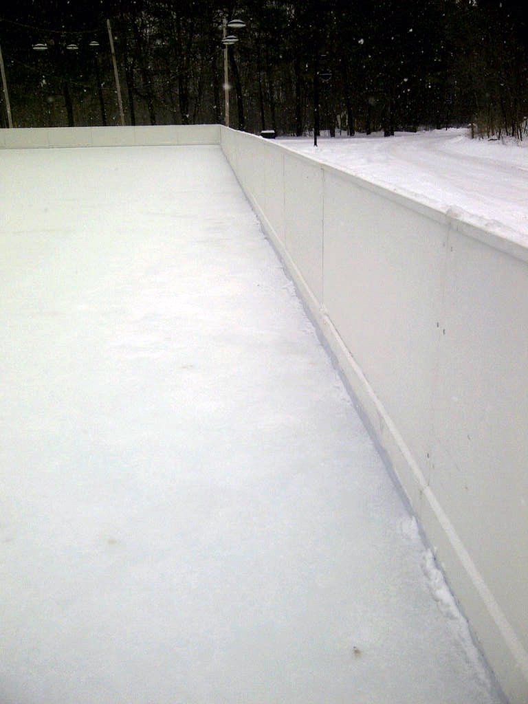 A snowy ice rink with a white fence and trees in the background.