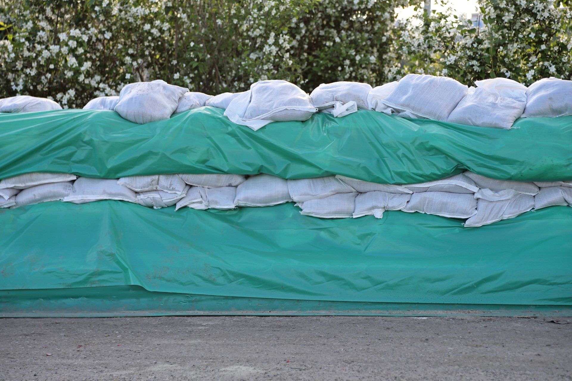 Sandbags behind a green tarp, providing a barrier, in front of bushes. Cover-Tech Emergency Relief Products.