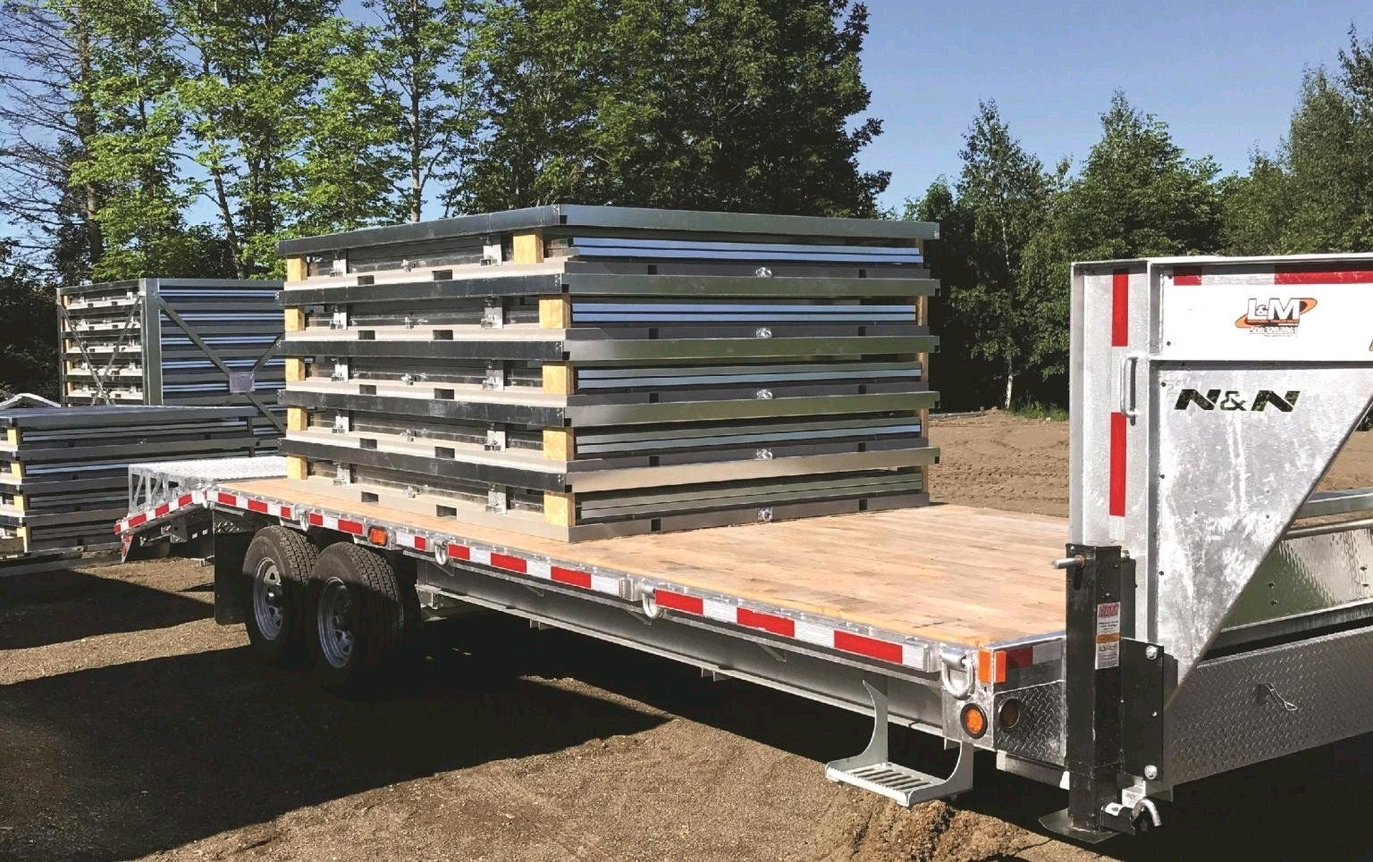 Flatbed trailer loaded with stacked metal panels, another similar stack in the background, outdoors on a sunny day. Cover-Tech Portable Containers.