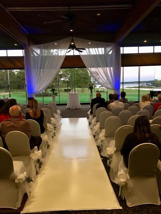 Wedding ceremony setup: white aisle runner, chairs with floral accents, sheer curtains, view of greenery.