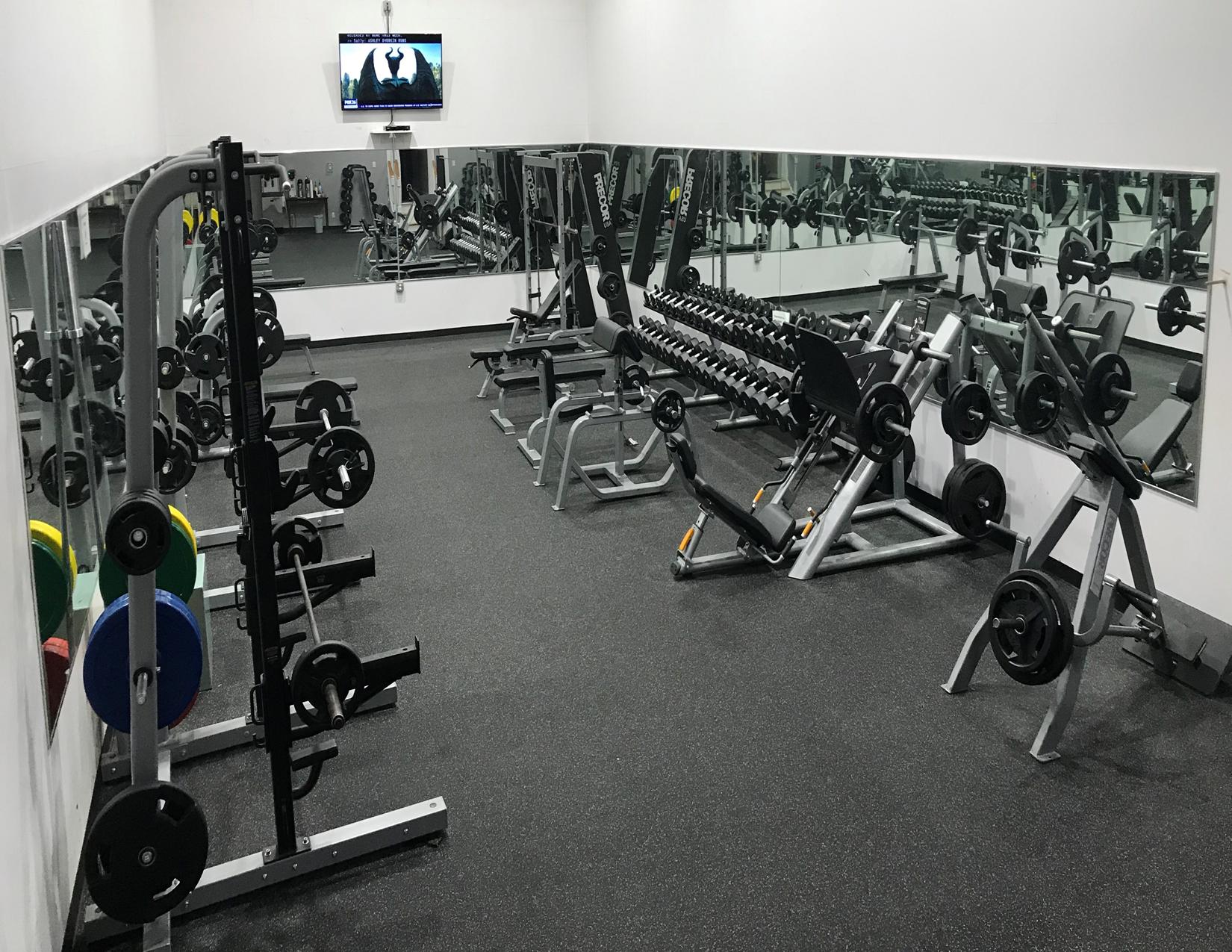 Gym interior with weight racks, dumbbells, and mirrors. Dark floor. TV mounted on the wall.
