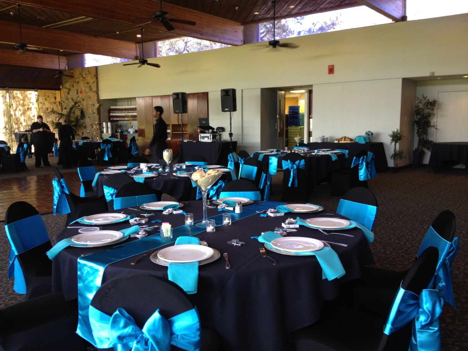 Wedding reception setup: Round tables with black tablecloths and teal accents; venue with large windows.