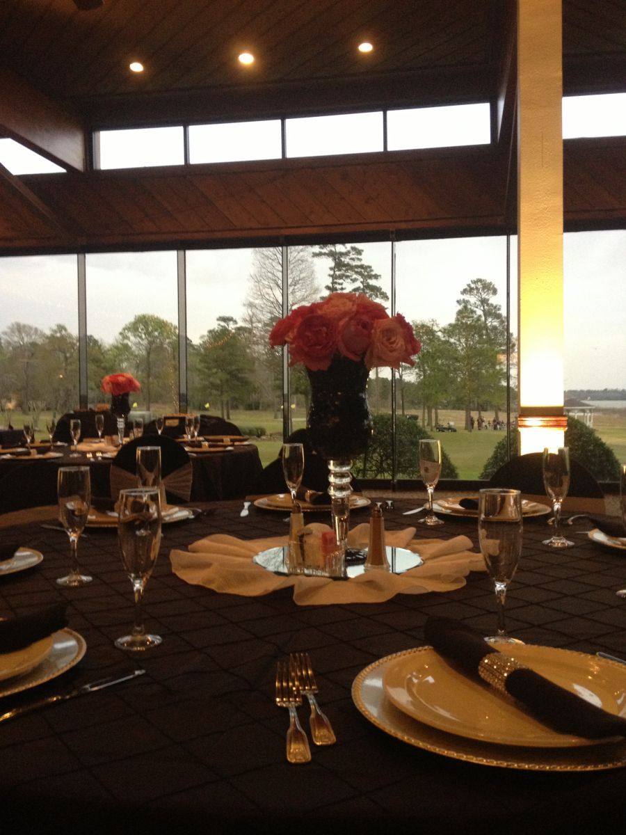Formal table setting with pink flower centerpiece, black tablecloth, and view of trees outside windows.