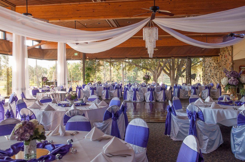 Wedding reception hall decorated with white and purple linens, draped fabric, and a chandelier.