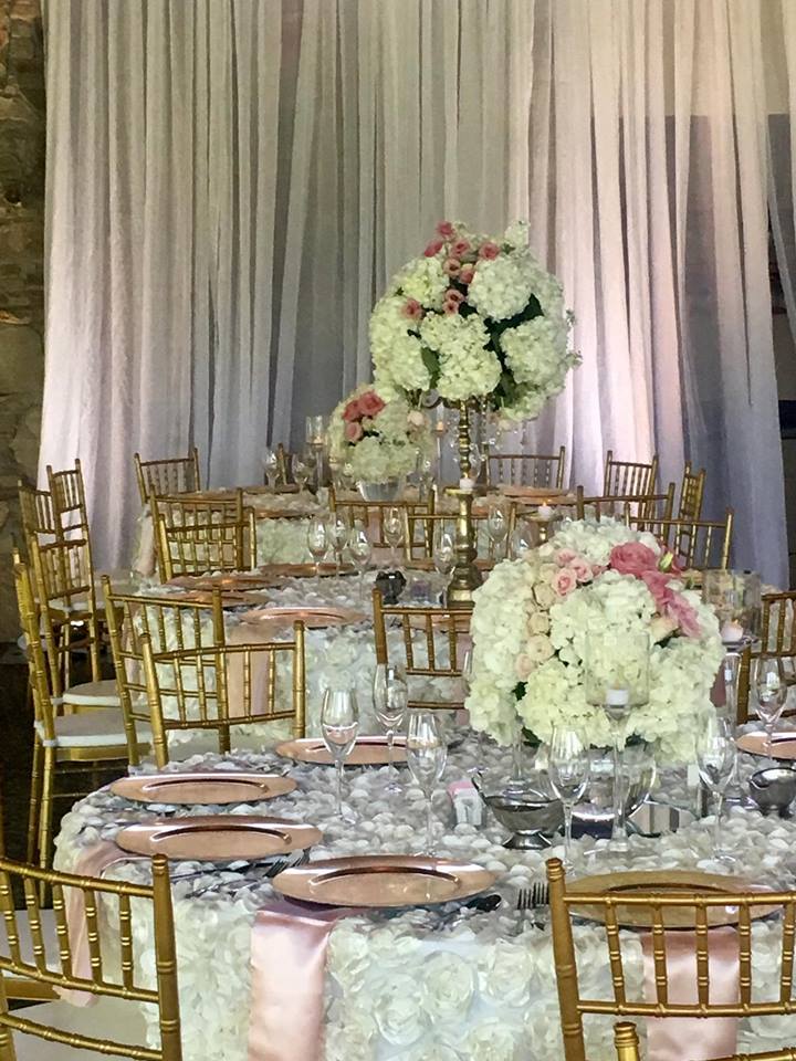 Elegant wedding reception table setting with floral centerpieces, gold chairs, and sheer backdrop.