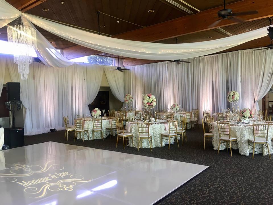 Wedding reception hall with white draped fabric, round tables with gold chairs, and a dance floor.