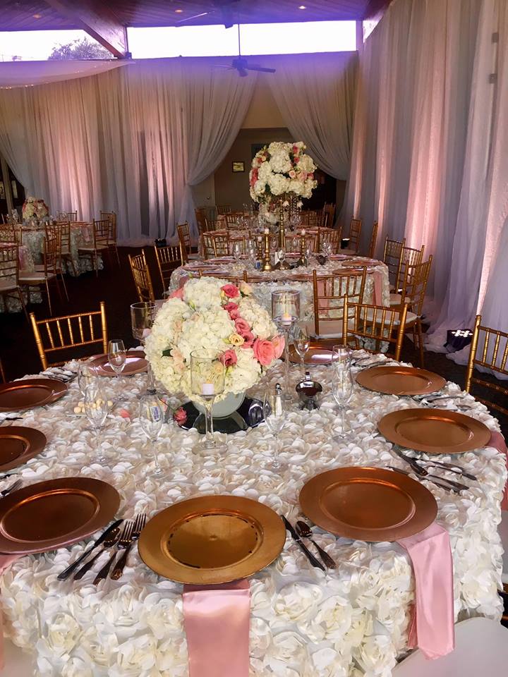 Elegant event space set with round tables, floral centerpieces, gold chairs, and rose-colored accents.