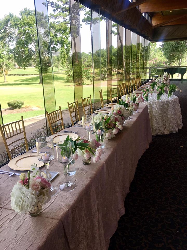 Long banquet table set with flowers, gold chairs, and a window view of green lawns.
