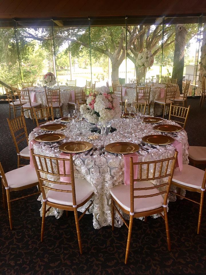Round table set for a formal event, gold chairs, lace tablecloth, pink runner, floral centerpiece, outdoor setting.