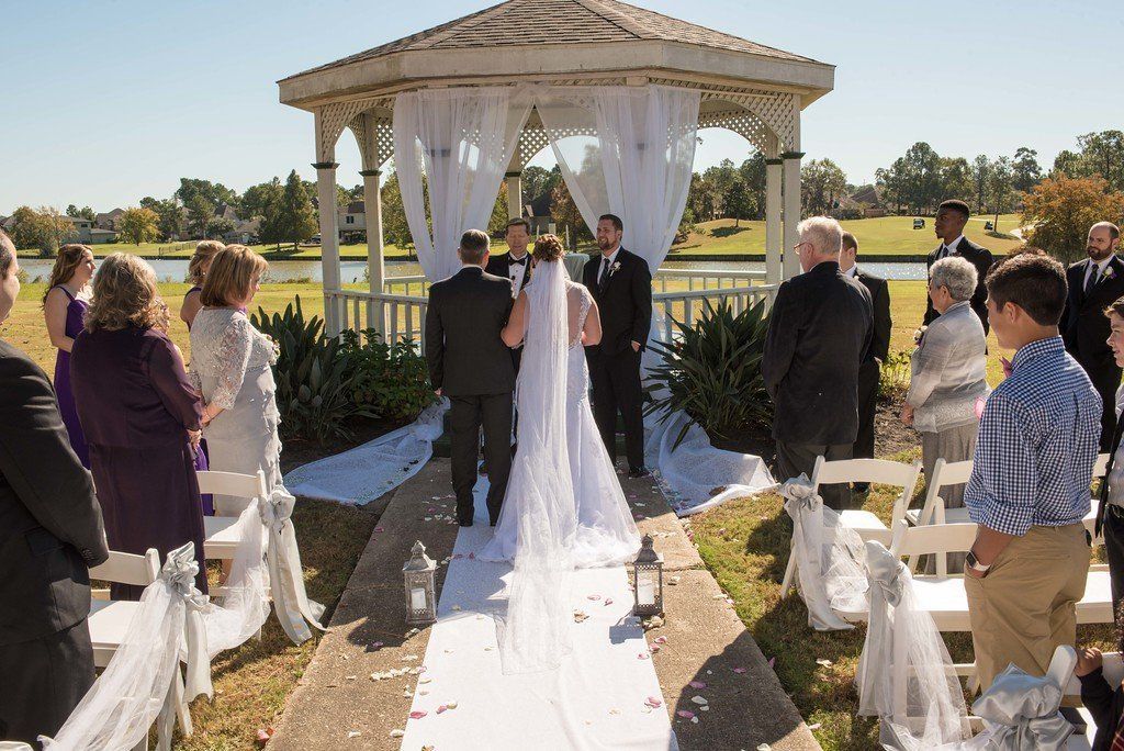 Wedding ceremony by a lake: couple at altar, guests watch. White aisle, gazebo, sunny day.