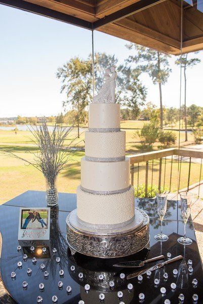 Five-tier wedding cake with silver accents, atop a shiny black table with champagne glasses and a scenic outdoor backdrop.