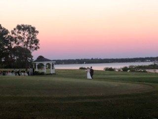 Wedding couple walks on a golf course towards a lake at sunset. A gazebo and guests are visible.