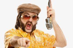 A man with a beard is holding a microphone and pointing at the camera.