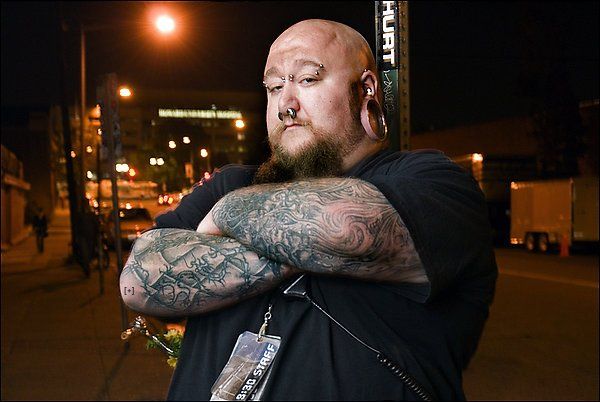 Man with tattoos and facial piercings stands outdoors at night, arms crossed, with a serious expression.