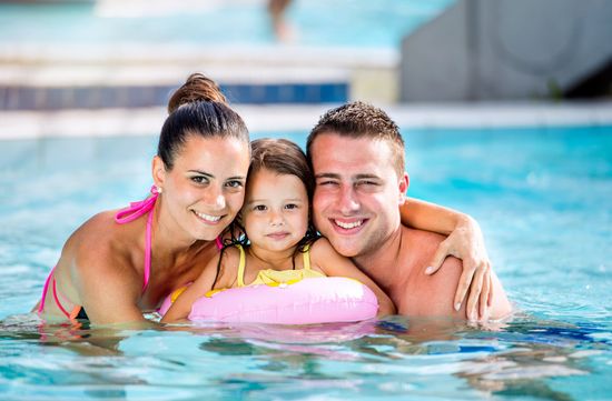 Family in a swimming pool smiles at the camera; mother in pink, daughter with floaty, father with arm around.