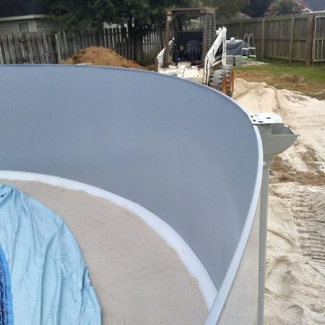 Construction of a swimming pool; curved concrete edge with gray interior, tan sand base, and construction equipment in the background.