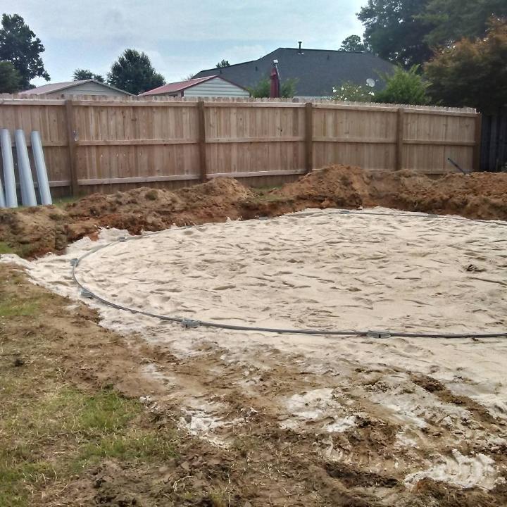 A circular area of sand and dirt is prepared for a pool. Fencing and pipes are in the background.