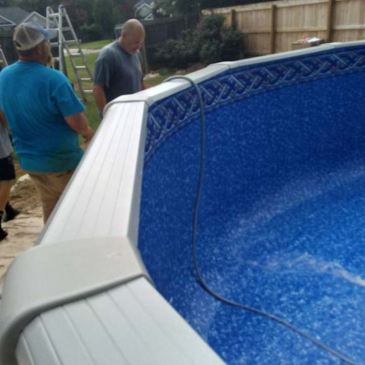 Men inspecting a partially filled, blue-lined above-ground pool. Beige top border, green yard, and wooden fence visible.