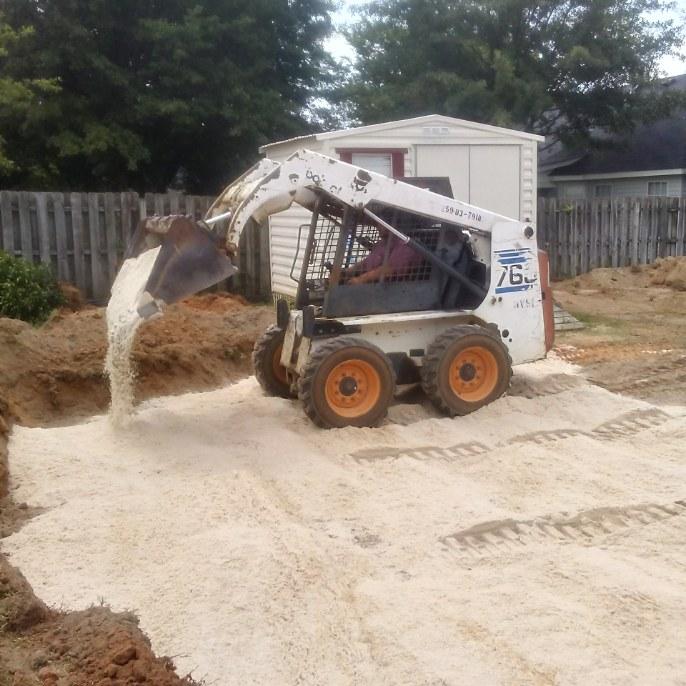 Bobcat skid steer spreading sand in a backyard excavation near a fence and shed.