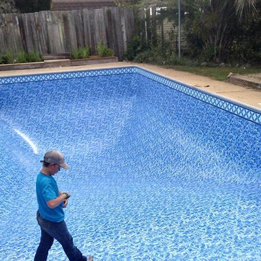Person walking in an empty blue-tiled swimming pool. The pool is in a yard with a wooden fence.