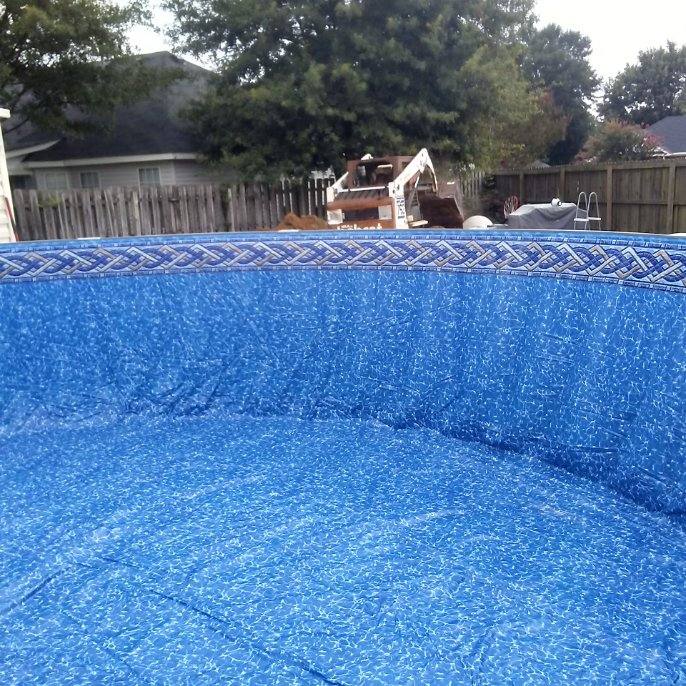 Blue, patterned pool liner, partially installed; backhoe visible in background near wooden fences.