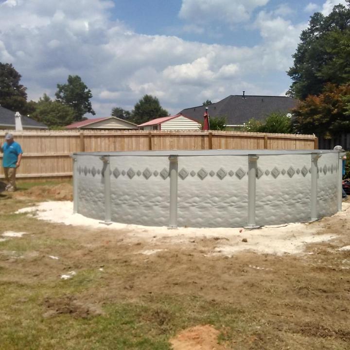 Above-ground pool installation in a backyard, surrounded by dirt. A person stands near the pool.