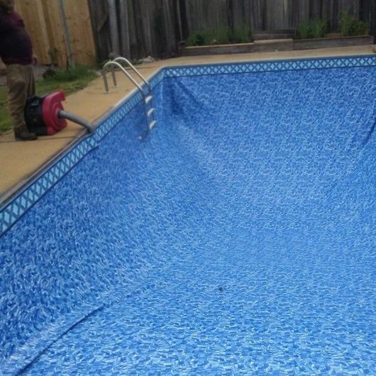 A worker uses a blower to install a blue patterned vinyl pool liner in an outdoor pool.