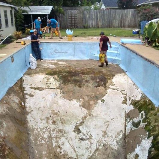 Workers repairing a drained, empty swimming pool. Concrete and blue paint visible.