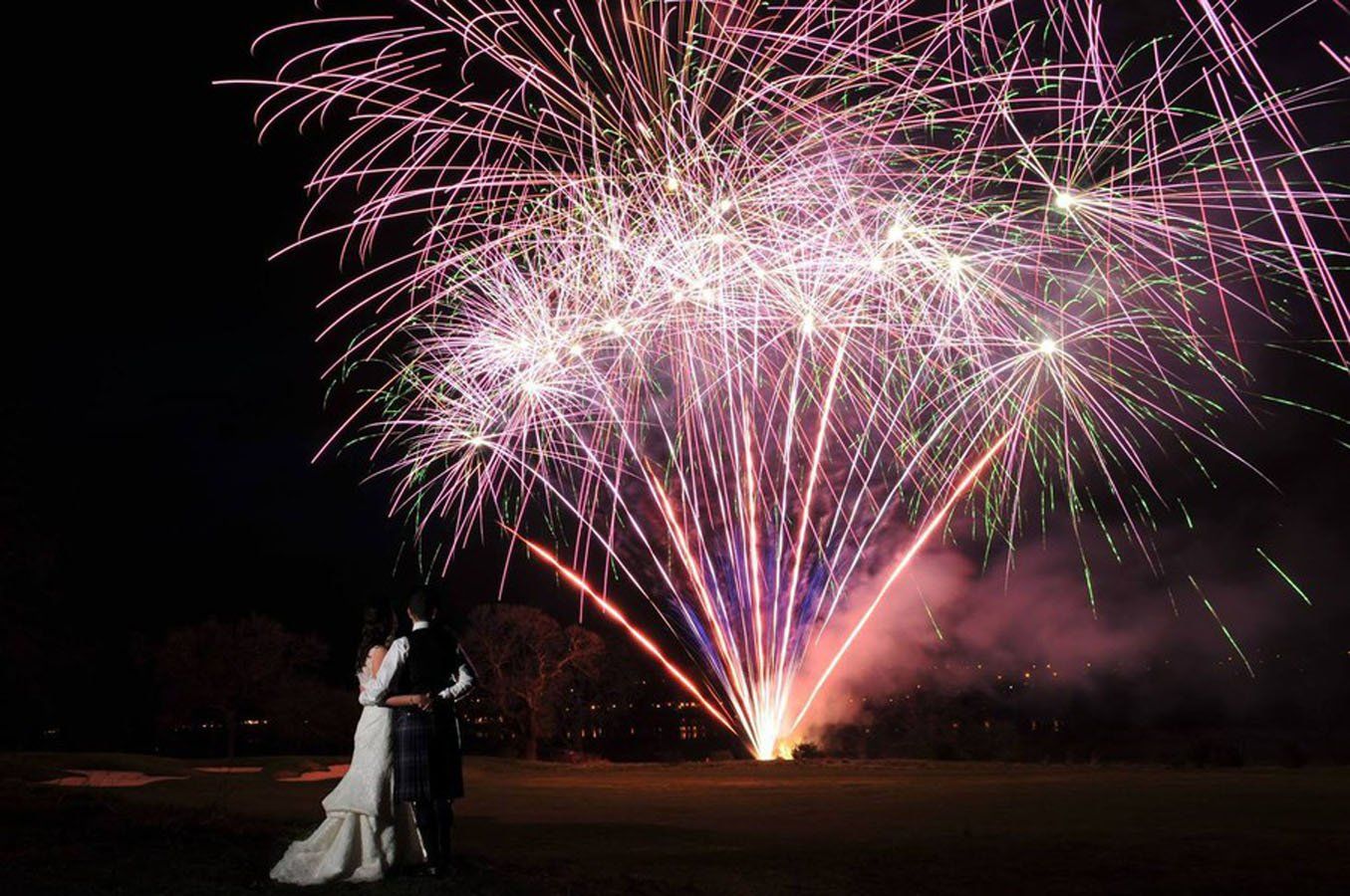 Una coppia si abbraccia, in controluce, mentre guarda i fuochi d'artificio che esplodono nel cielo notturno con i loro colori rosa e bianchi.