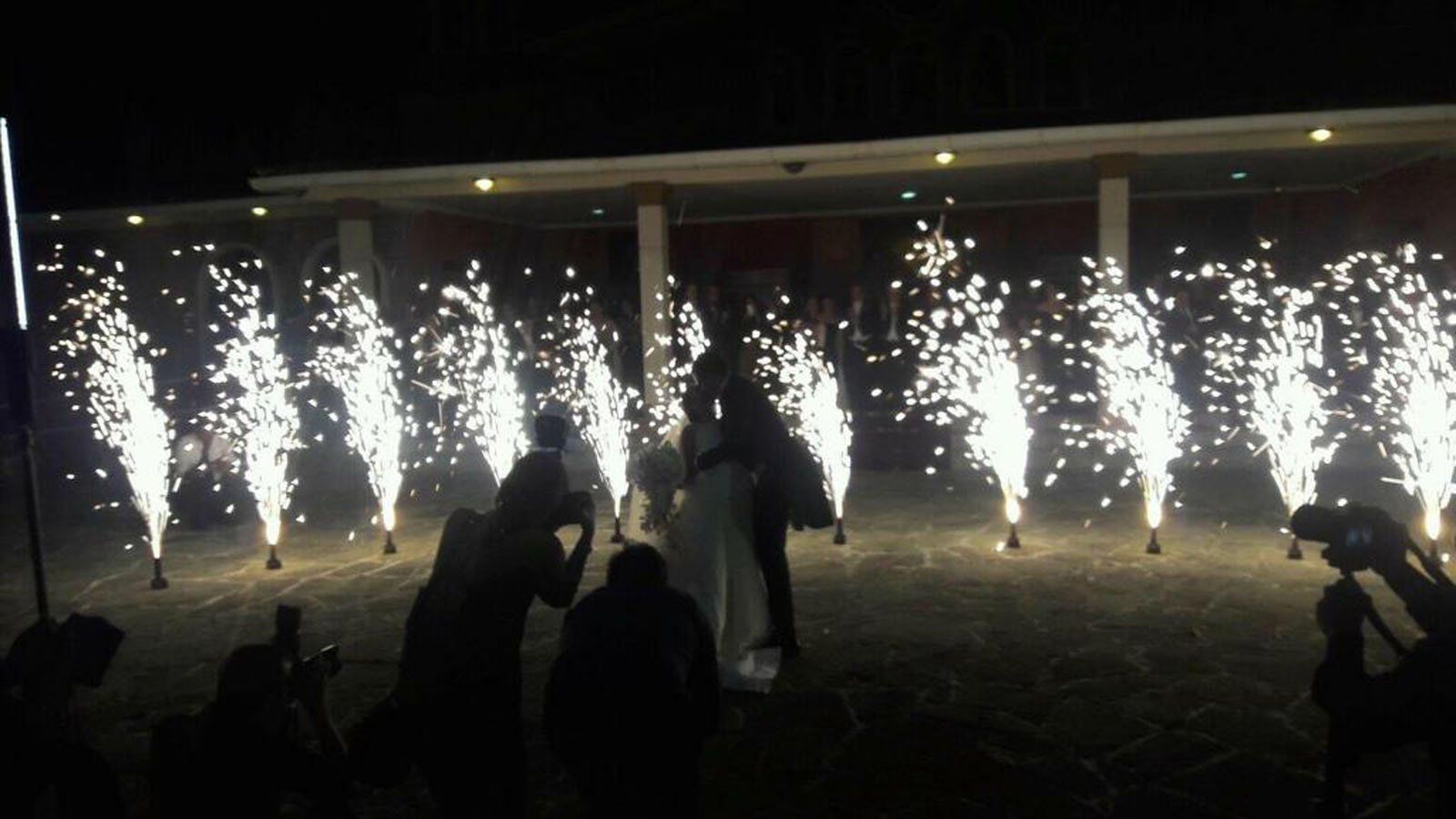 Una coppia si bacia tra fontane di scintille di notte durante una festa, circondata da spettatori con telecamere.