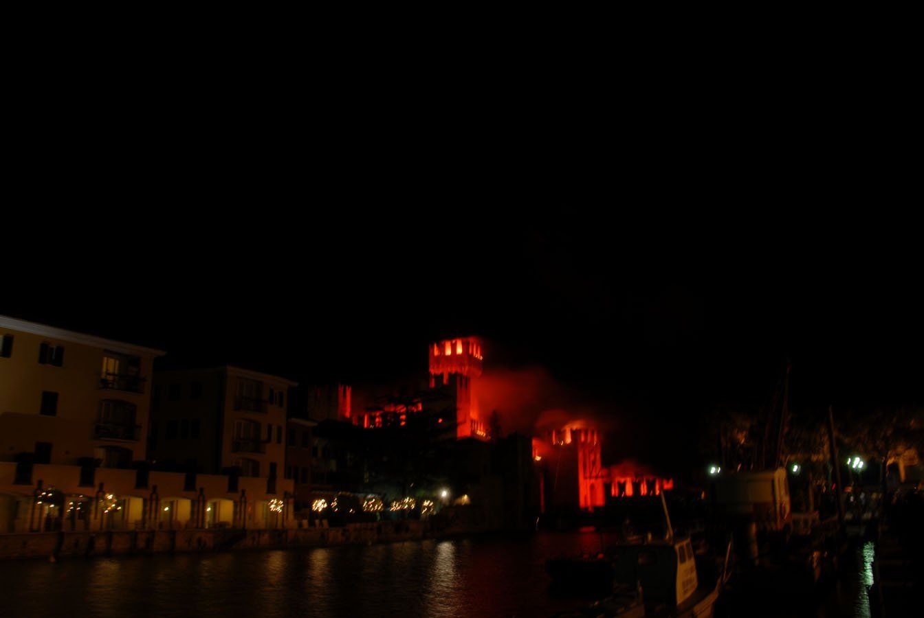 Castello illuminato di notte, luci rosse sulla struttura, acqua scura in primo piano, edifici lungo la riva.