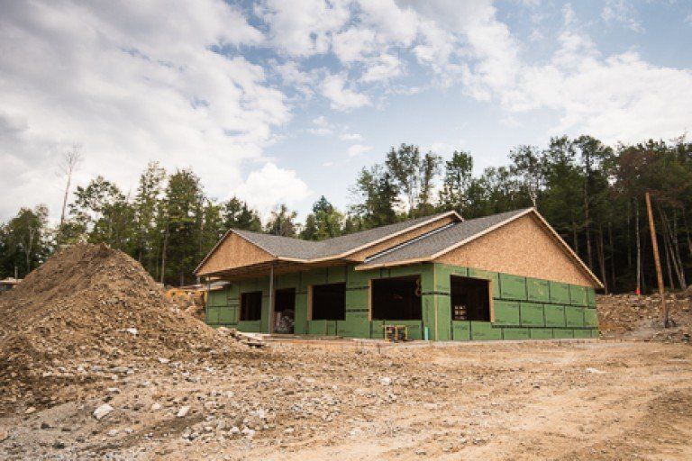A house under construction with exposed wood framing, green sheathing, and a dirt pile in front.