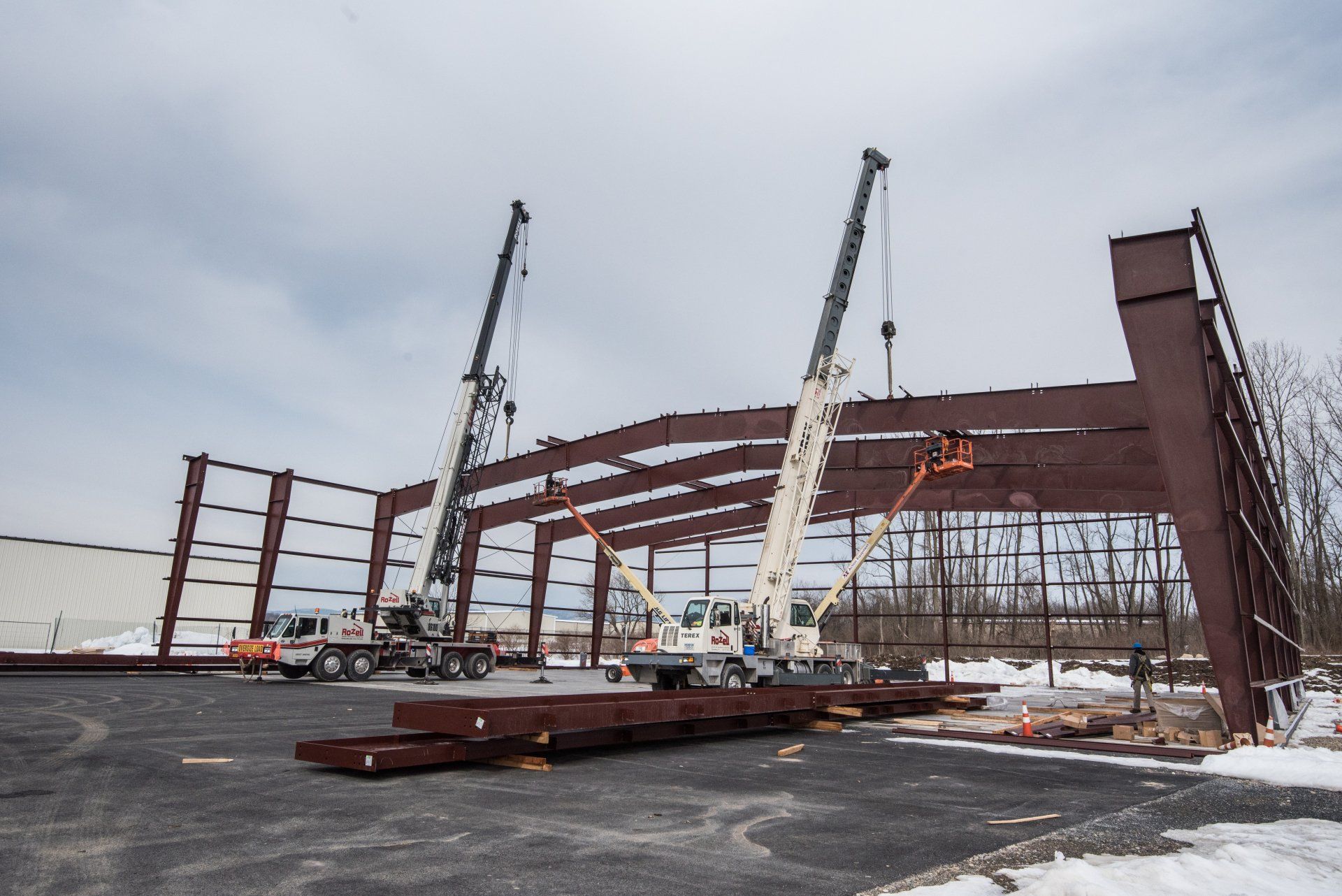 Two cranes lifting steel beams, constructing a building frame outdoors on a snowy day.