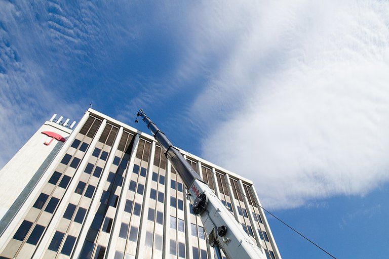 Crane reaching towards a tall white building with dark windows under a blue sky with fluffy clouds.
