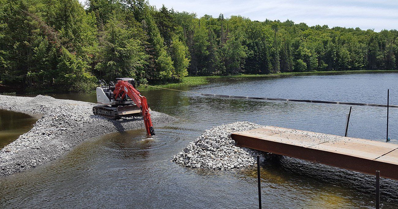 An excavator placing rocks in a lake to create a shoreline; trees and a dock are in the background.