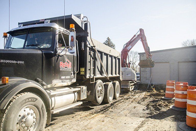 Black dump truck beside excavator at a construction site, orange traffic barrels present.