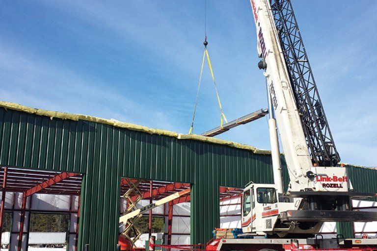 Construction crane lifting a metal beam onto a green metal building under a blue sky.