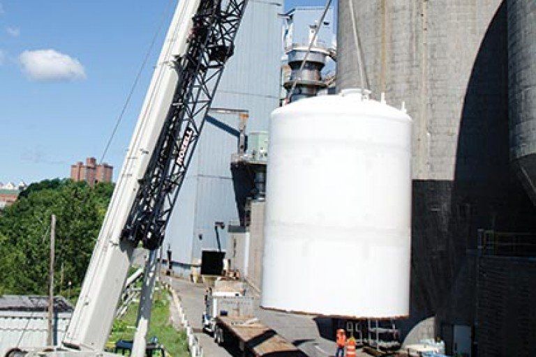 A crane lifts a large white cylindrical tank at an industrial facility, blue sky, concrete silos in the background.